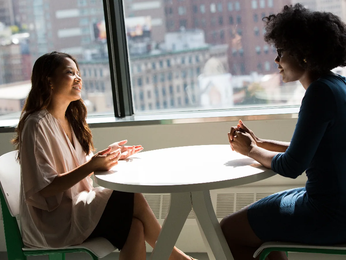 Two colleagues in conversation across a table in a bright office overlooking the city.